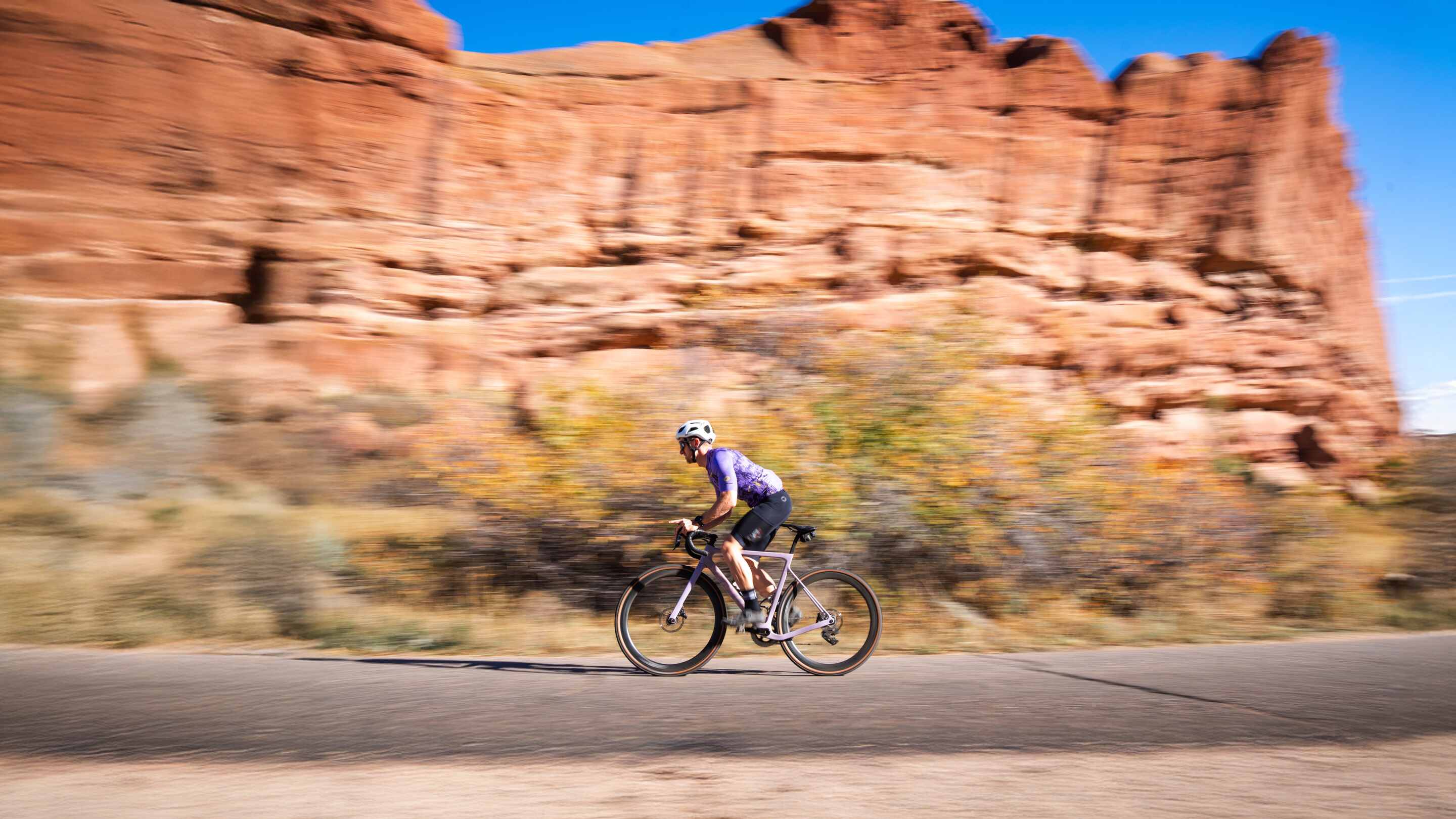 Cyclist racing up road with Red Rocks in Morrison Colorado behind him while wearing a purple cycling jersey and black cycling cargo bib shorts