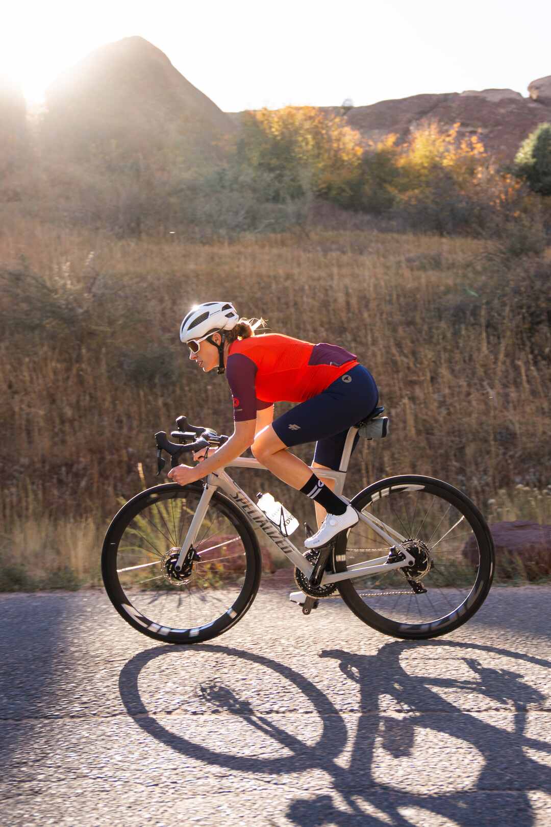 Female cyclist wearing orange cycling jersey and blue cycling bibs with sun setting behind her