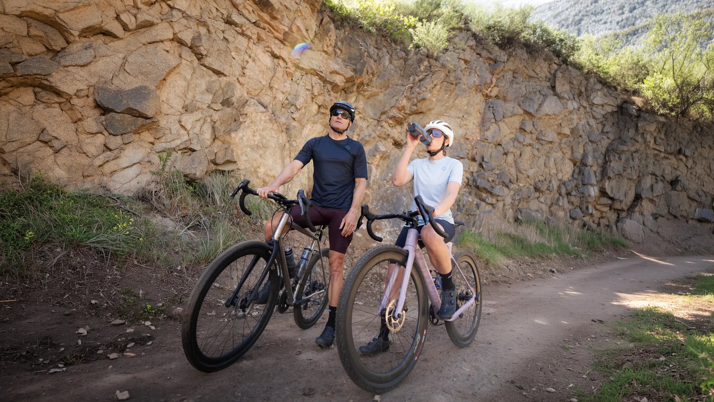 Two cyclists on a dirt path, one drinking from a water bottle