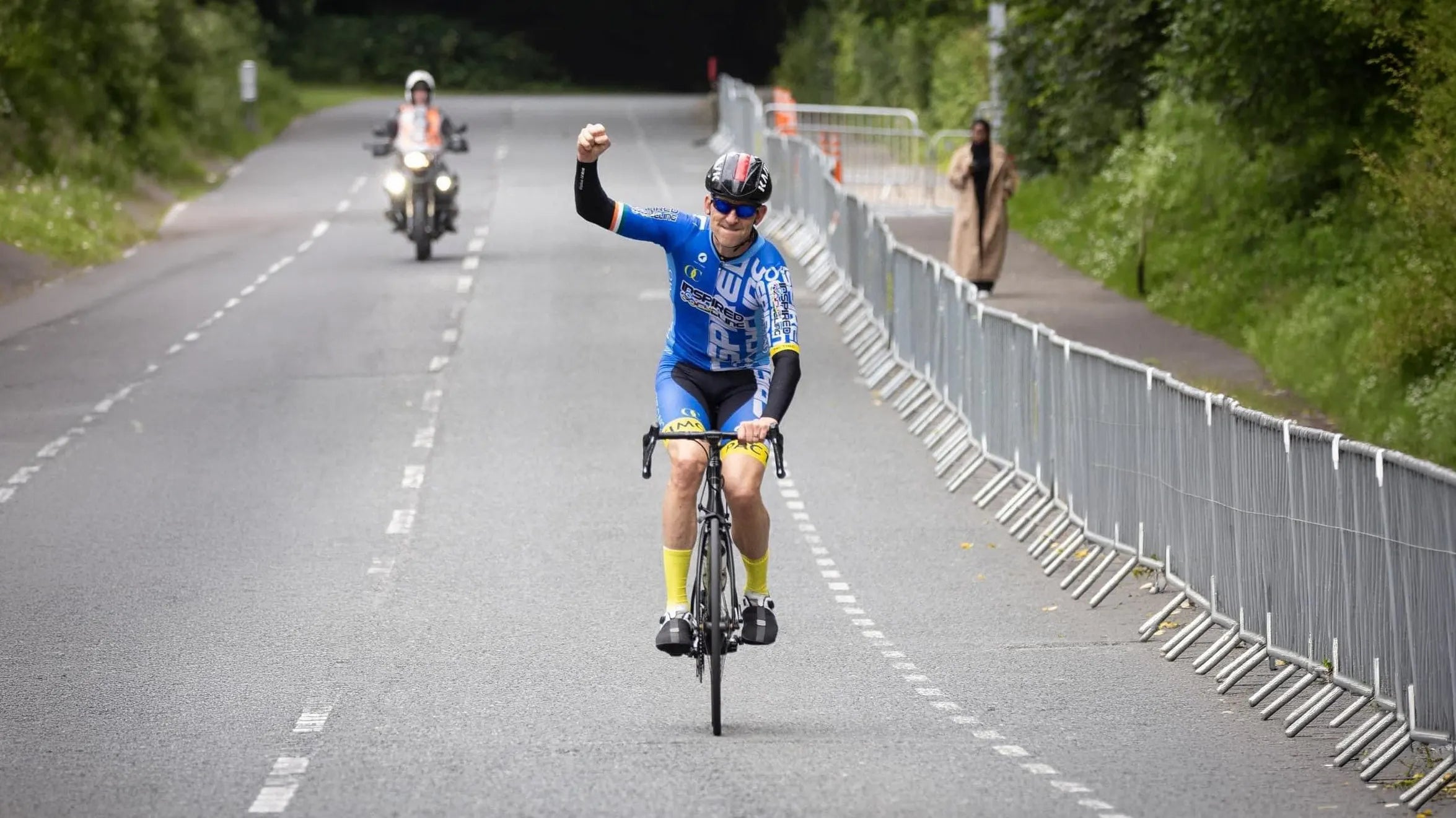 A cyclist in a blue and white jersey rides a road bike down a paved road with a raised fist, with a motorcycle and barriers in the background.