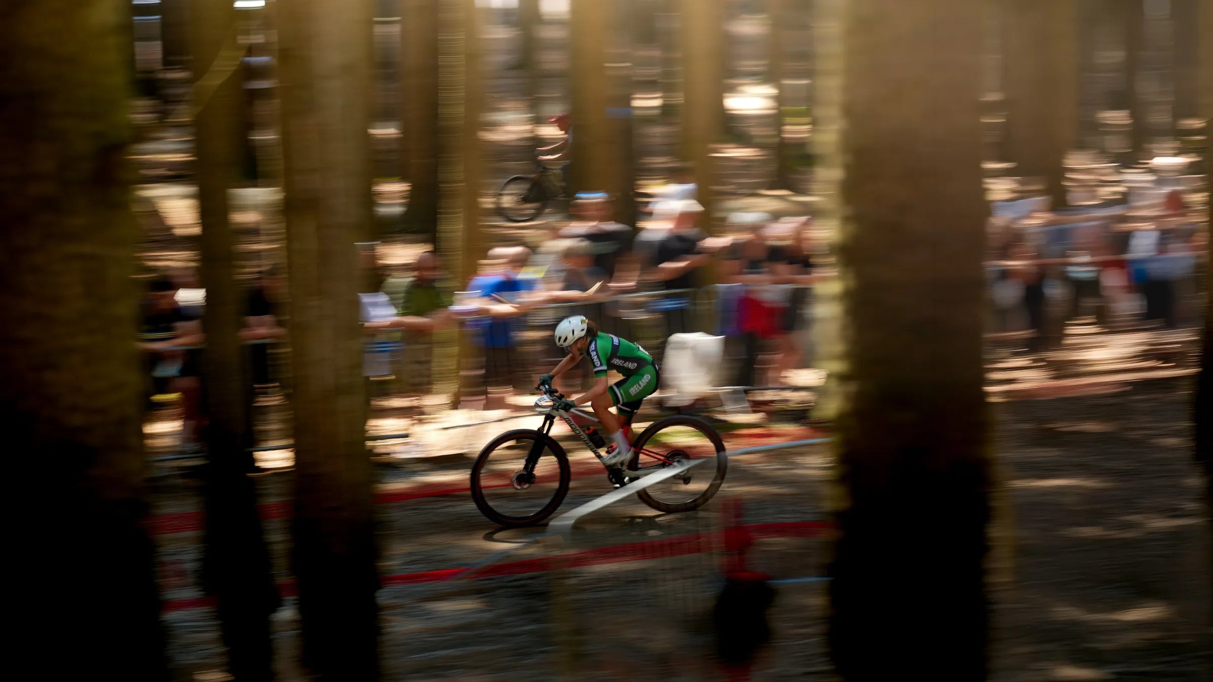 A mountain biker in a green and black cycling kit rides through a dense forest during a race, with blurred spectators and tree trunks in the background.
