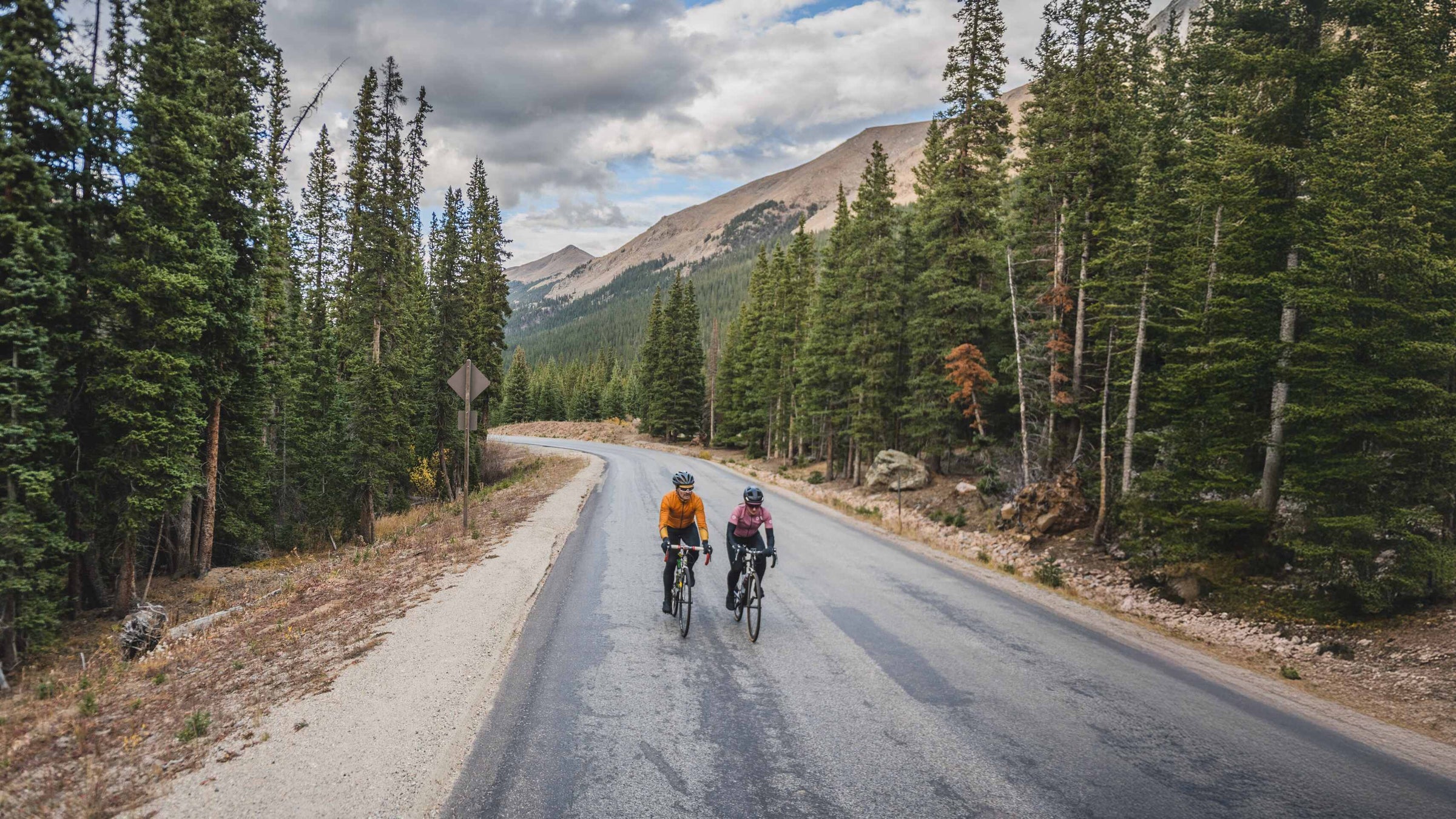 Two cyclists on a road surrounded by trees and mountains. Black Friday Cycling clothing sale