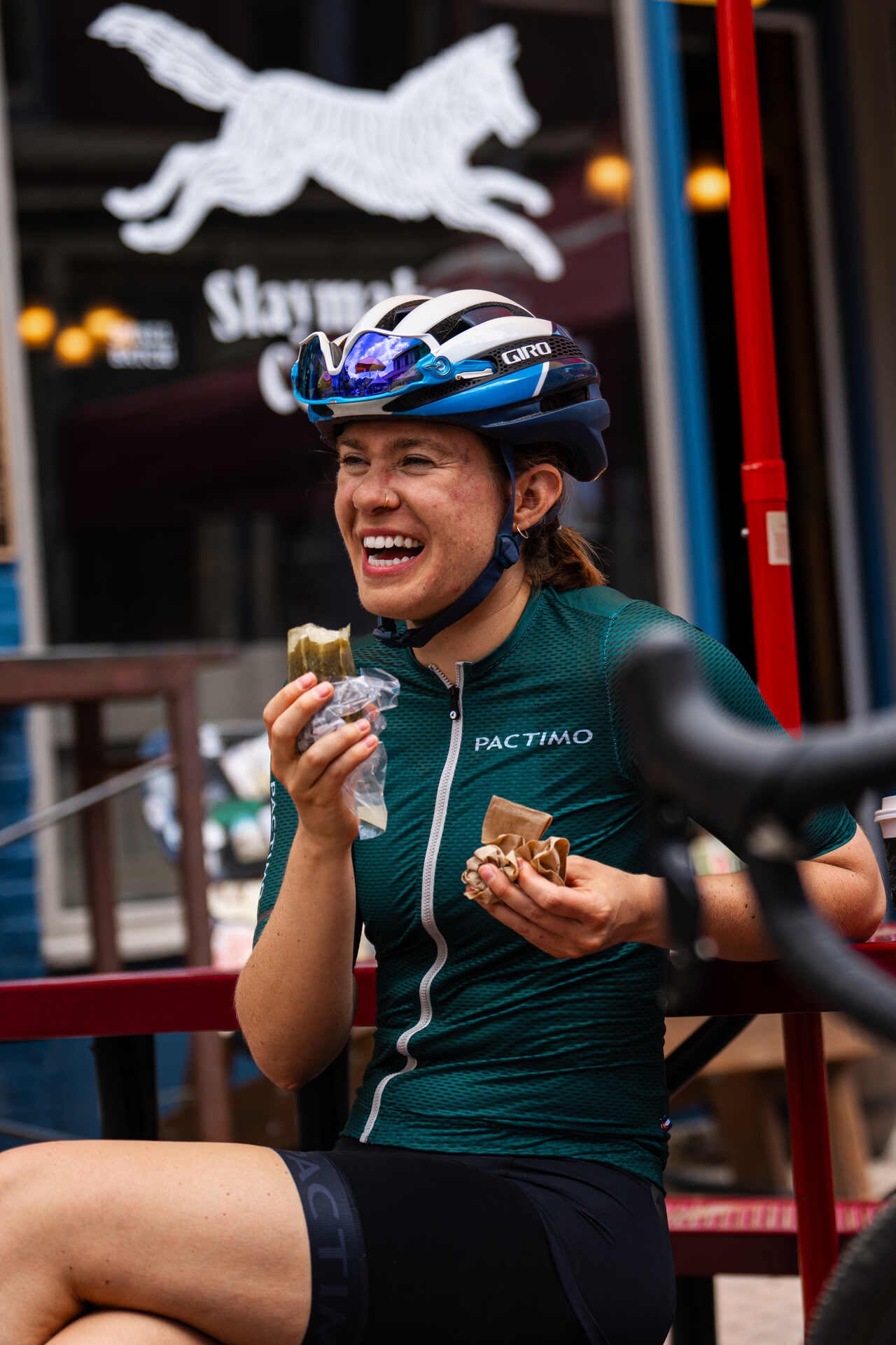 A smiling woman in a Pactimo jersey taking a snack break outside a café after a ride