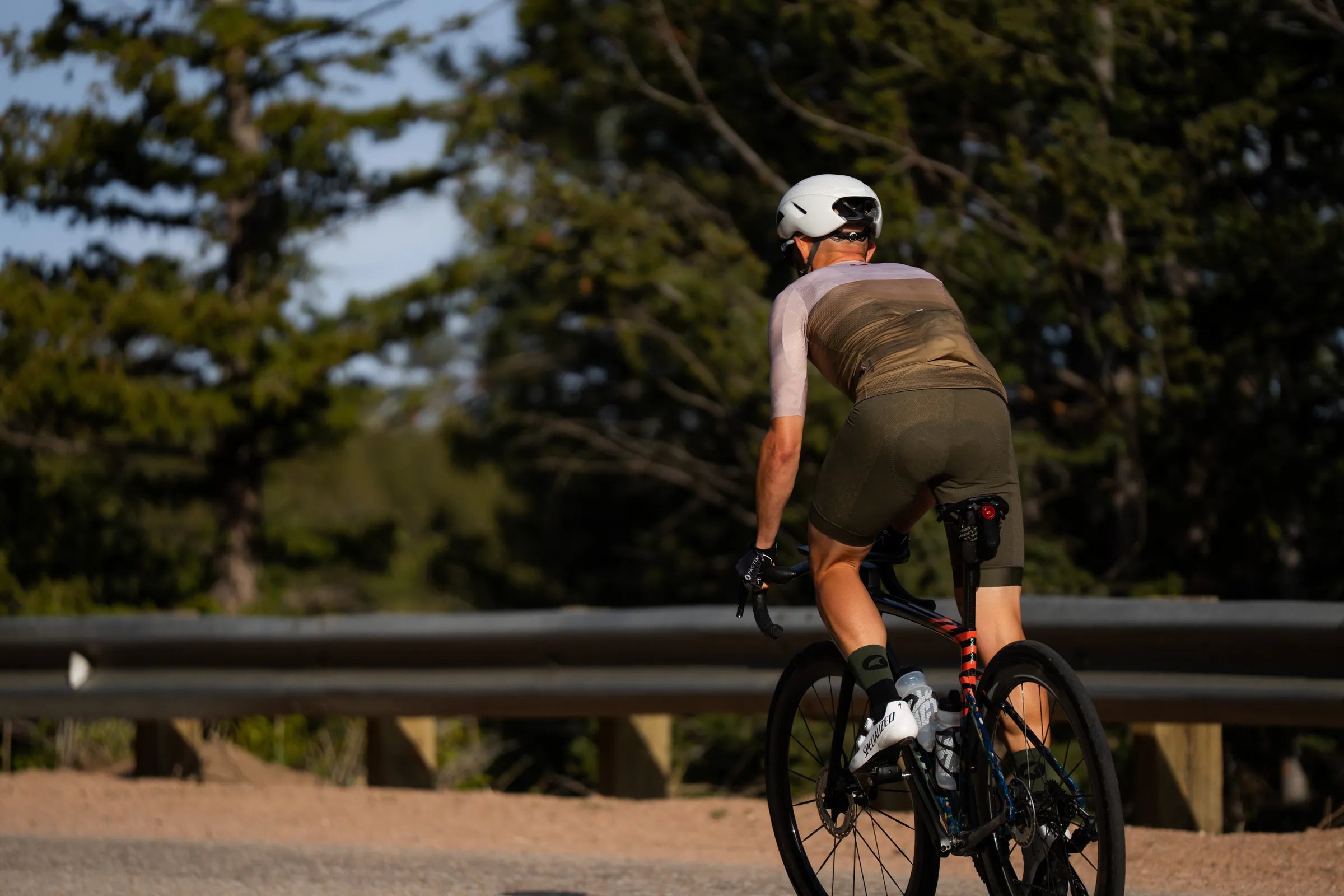 A cyclist wearing a white helmet, a beige and olive green cycling jersey, and dark green shorts rides a road bike on a dirt road with trees and a guardrail in the background.