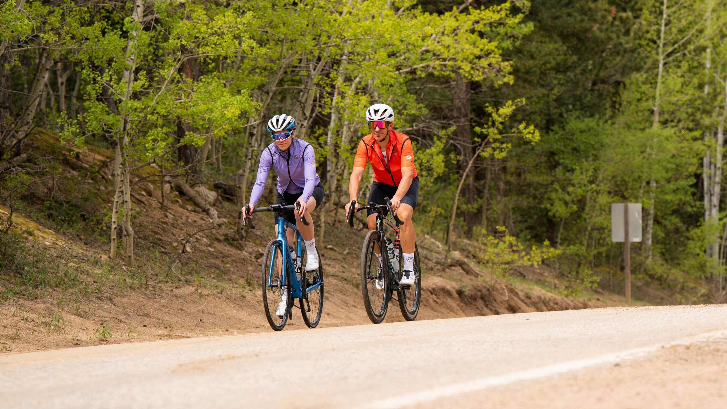 Two cyclists on a road surrounded by trees in Pactimo Divide cycling wind vests