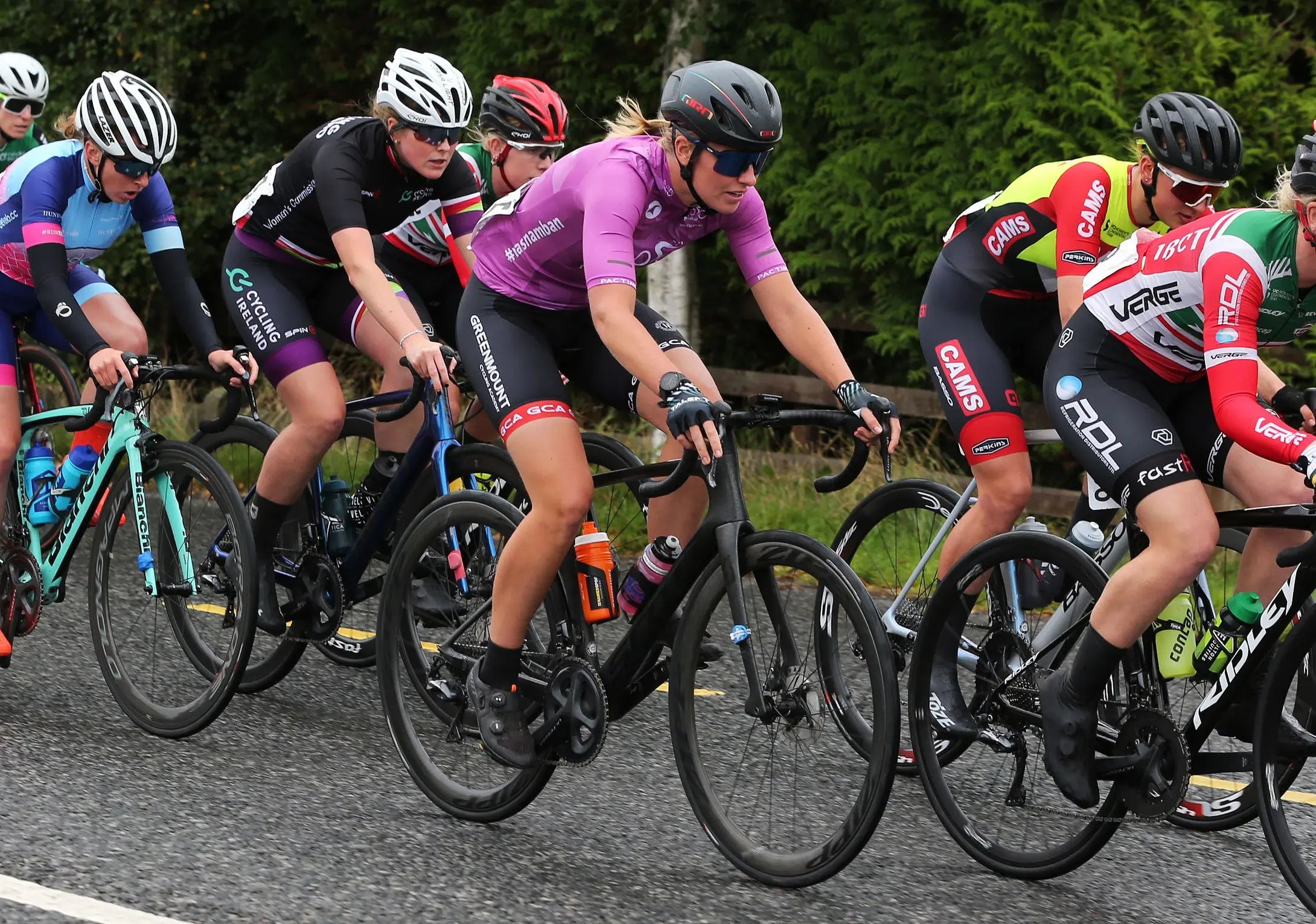 A group of professional road cyclists riding closely together on a paved road during a race. The central rider wears a bright purple jersey and black shorts, with other riders in multicolored team kits visible around her.