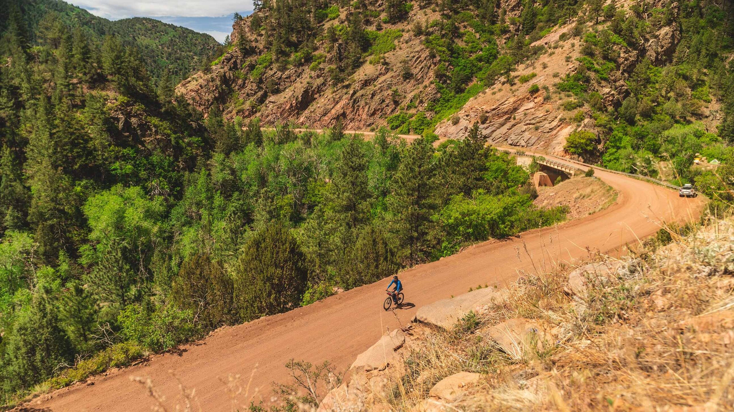 Cyclist on a dirt road surrounded by green trees and mountains